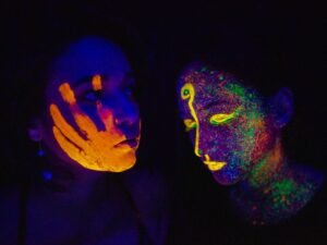 Colorful face paint glows under UV light, showcasing unique designs on two women's faces.